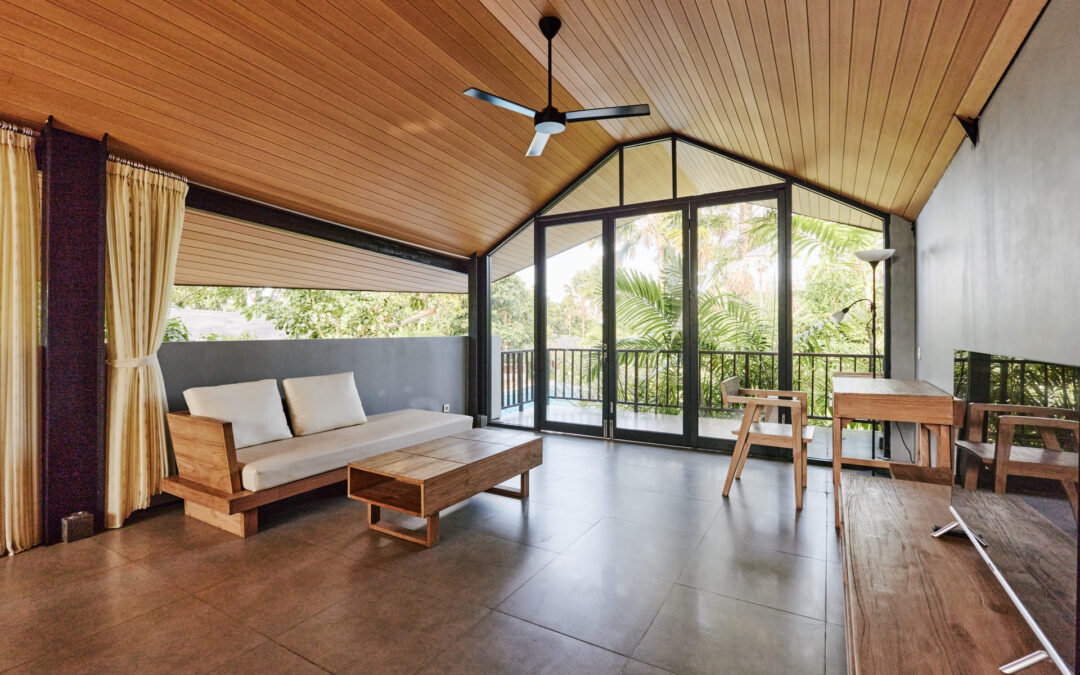Vista interna di una moderna veranda in legno e vetro chiusa per casa. L'ambiente luminoso presenta un alto soffitto inclinato con perline di legno e un ventilatore centrale. Le ampie pareti vetrate con infissi scuri mostrano un balcone con vista su alberi verdi rigogliosi. L'arredamento in legno coordinato include un divano con cuscini bianchi, un tavolino da caffè, una scrivania con sedia e un mobile TV. Il pavimento è rivestito con piastrelle color terracotta sfumato.